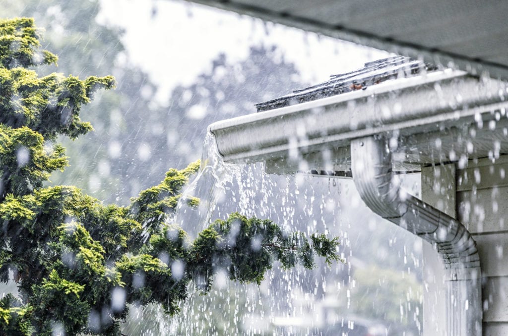 Drenching downpour rain storm water is overflowing off the tile shingle roof - streaming, rushing and splashing out over the overhanging eaves trough aluminum roof gutter system.
