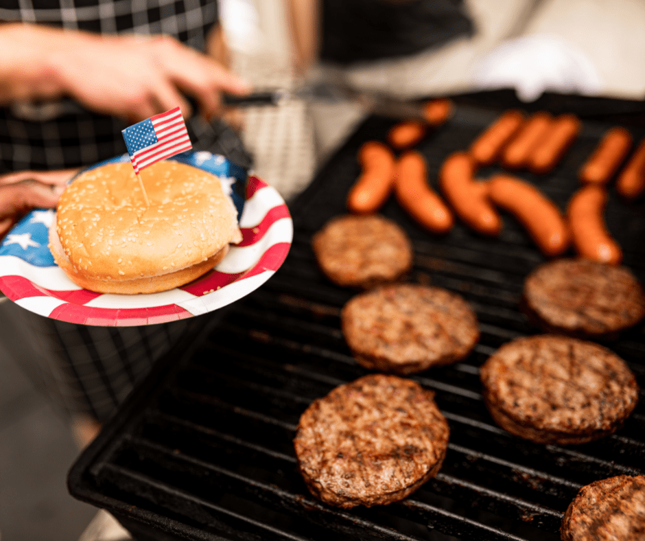 Burgers and hotdogs on a grill. In the forgound, a burger bun with an American flag sticking in the top.