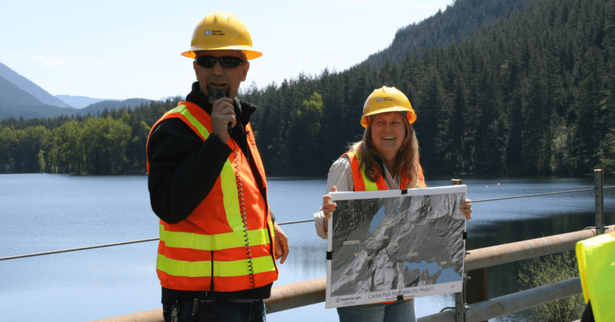 Two people speaking at a lake, holding a map of the Cedar Falls Hydroelectric Project.